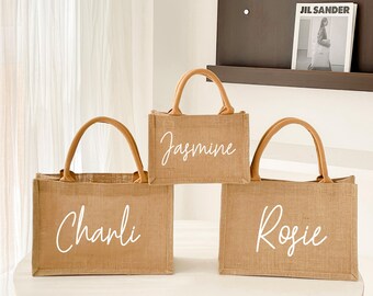 May include: Three rectangular jute tote bags with brown handles. Each bag has a white script name: "Charli", "Jasmine", and "Rosie". The bags are on a white surface.