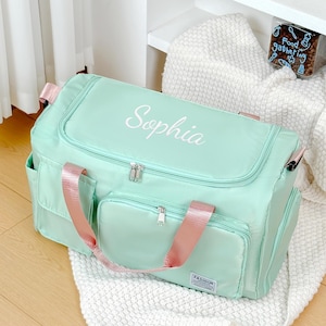 Op de afbeelding: Een mintgroene reistas met de naam "Sophia" in wit geborduurd. De tas heeft roze handvatten en accenten, met meerdere vakken met ritssluiting en een "Fashion" label.