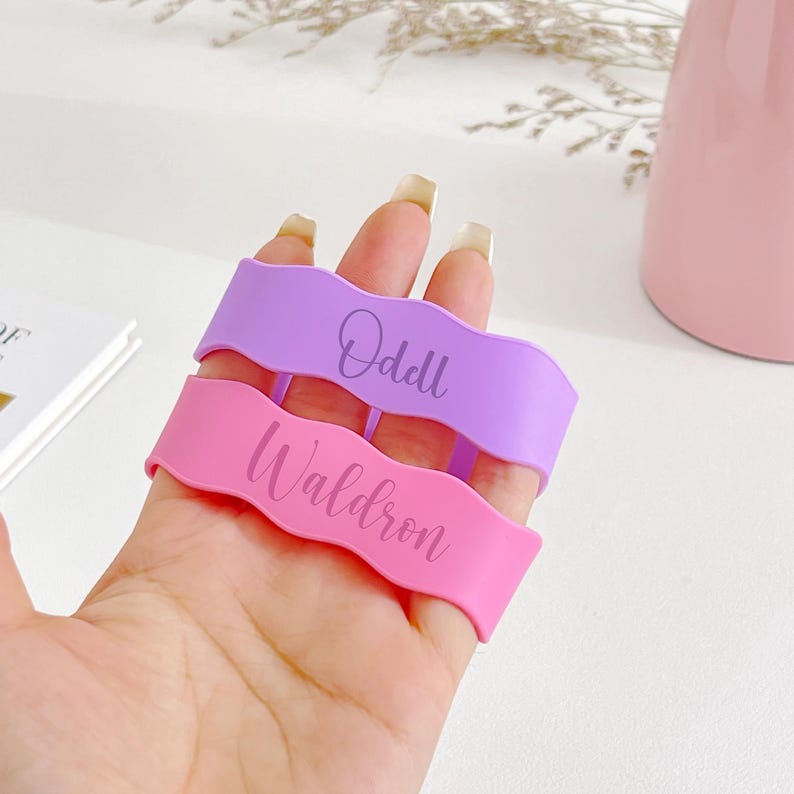 May include: Two wavy, silicone wristbands, one lavender and one pink, are held in a hand. The lavender band reads "Odell" and the pink band reads "Waldron" in a cursive font. The background includes a pink vase and a white book.