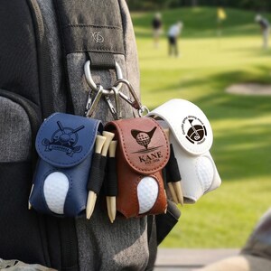 Könnte beinhalten: Drei Golfballhalter, die an einem Rucksack befestigt sind. Einer ist marineblau mit einem Golfschläger- und Ball-Design, ein anderer ist braun mit einem Golfball-Design und der dritte ist weiß mit einem Golfschläger- und Ball-Design. Jeder Halter hat einen Golfball und Tees.