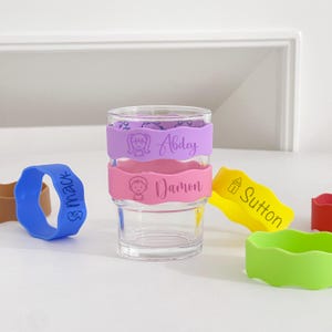 May include: A clear glass with two silicone bands in lavender and pink, each with a name and a cartoon illustration. Additional silicone bands in blue, yellow, green, and red are scattered around the glass. The bands are designed to fit around the glass.