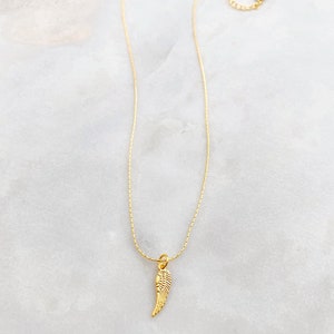 May include: A gold-coloured necklace with a small wing pendant. The pendant has detailed features, and the chain is adjustable. The necklace is shown on a white and grey marble surface.