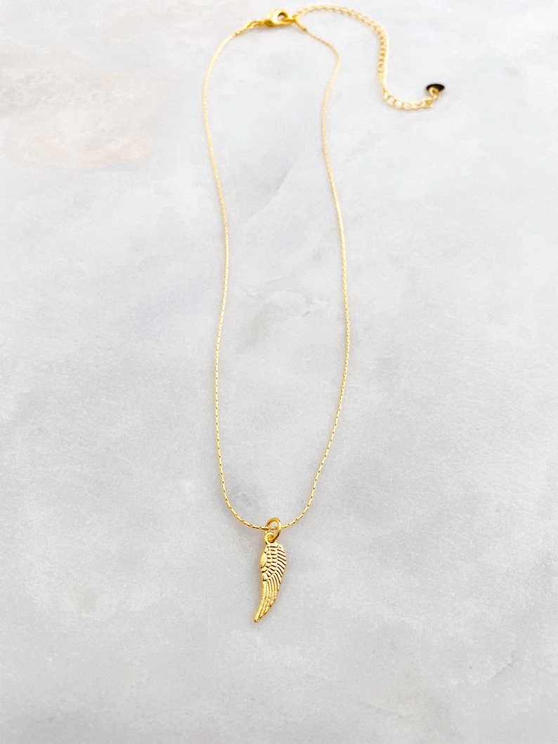 May include: A gold-coloured necklace with a small wing pendant. The pendant has detailed features, and the chain is adjustable. The necklace is shown on a white and grey marble surface.