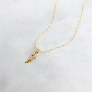 May include: A gold-coloured necklace featuring a small wing pendant. The wing pendant has feather-like detailing. The chain is thin and the pendant is centrally placed. The background is a white marble surface.