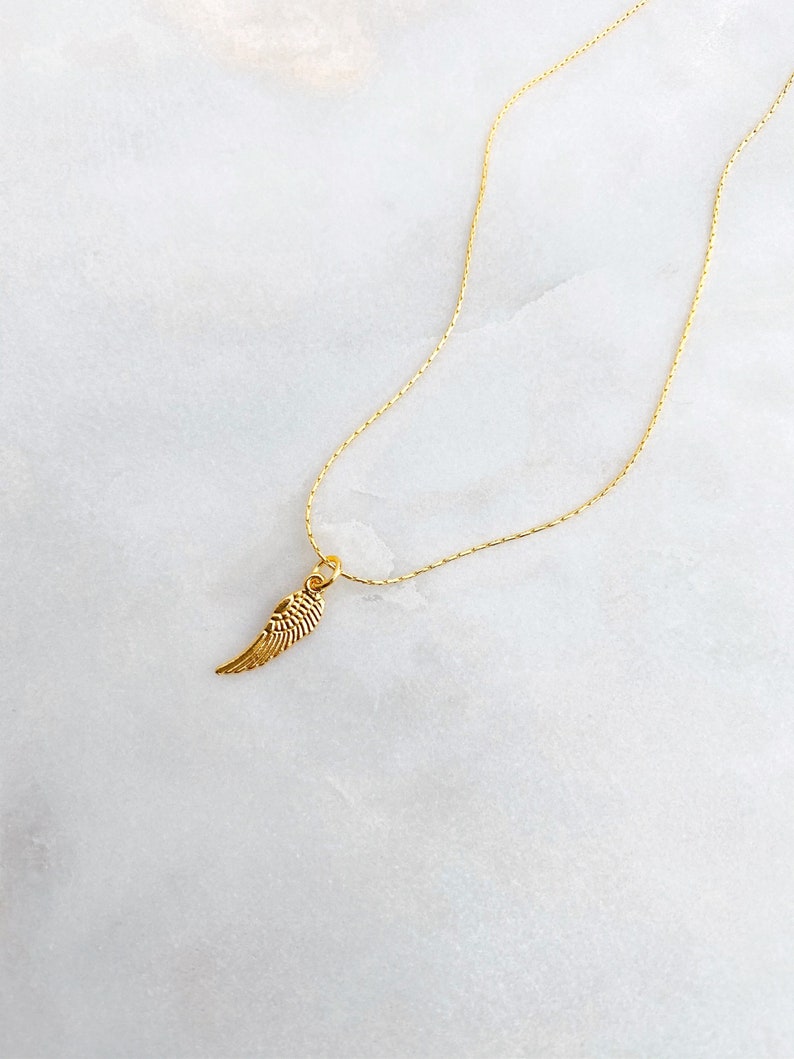 May include: A gold-coloured necklace featuring a small wing pendant. The wing pendant has feather-like detailing. The chain is thin and the pendant is centrally placed. The background is a white marble surface.