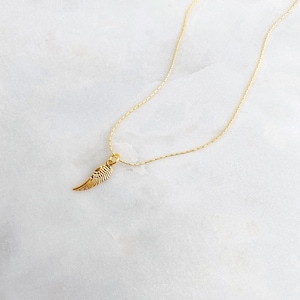 May include: A gold-coloured necklace with a small wing pendant. The wing is finely detailed, and the chain is slender and elegant. The necklace is displayed on a white and grey marble surface, creating a minimalist aesthetic.