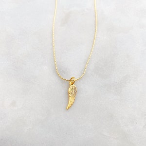 May include: A gold-coloured necklace with a small wing pendant. The pendant has detailed features, and the chain is thin and elegant. The necklace is shown against a light background, emphasising its simple beauty.