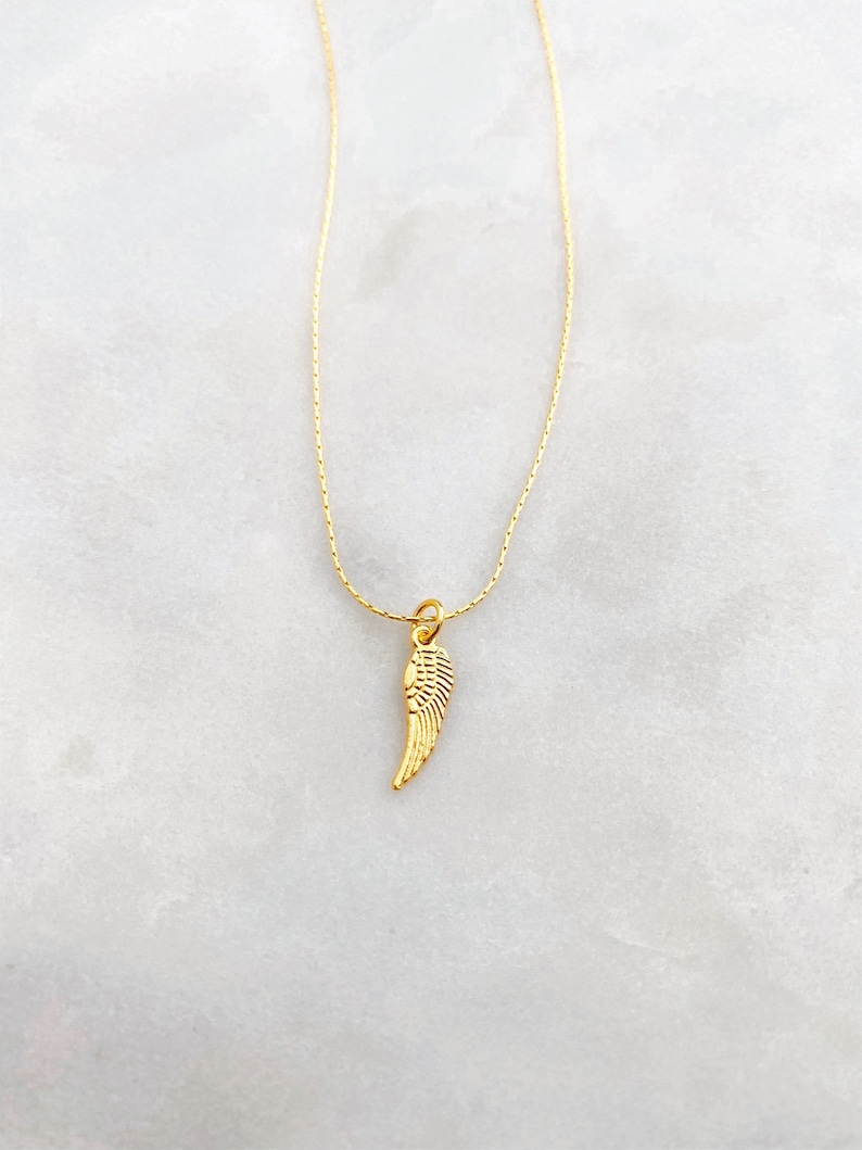 May include: A gold-coloured necklace with a small wing pendant. The pendant has detailed features, and the chain is thin and elegant. The necklace is shown against a light background, emphasising its simple beauty.