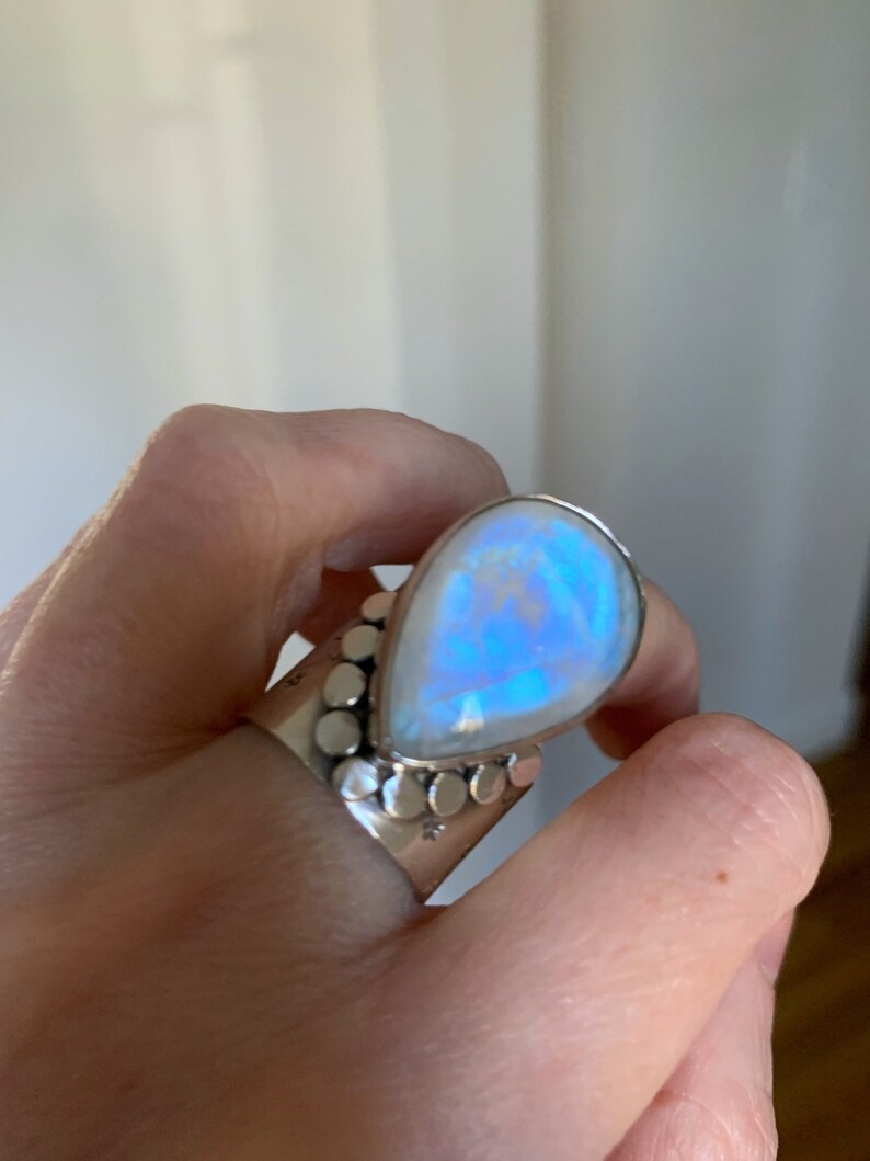 Pode incluir: Um anel de prata com uma grande pedra de lua em forma de l&aacute;grima. A pedra tem um brilho iridescente azul.