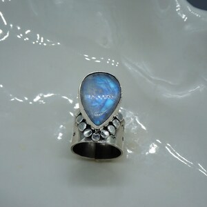 Pode incluir: Um anel de prata com uma pedra de lua azul em forma de l&aacute;grima engastada no centro. A banda &eacute; decorada com pequenas contas de prata.