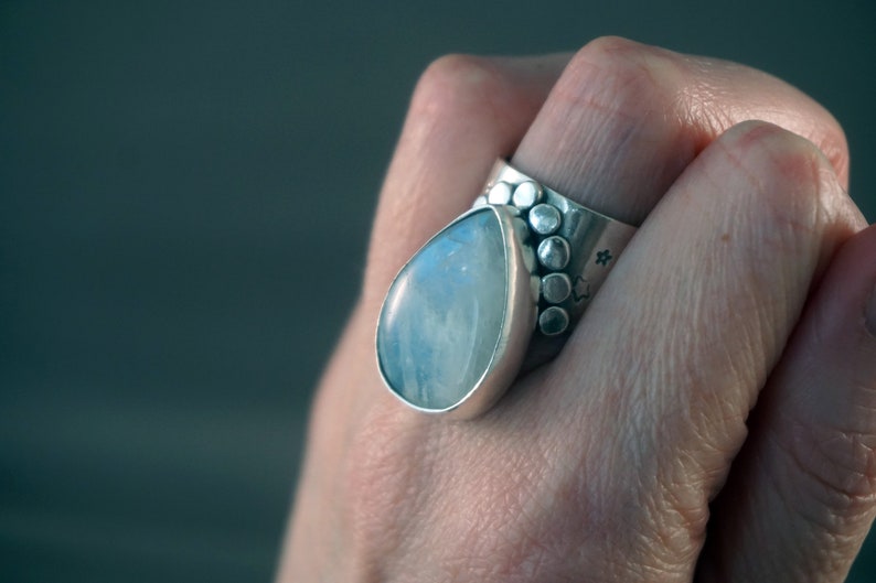 Pode incluir: Um anel de prata com uma grande pedra de lua em forma de l&aacute;grima engastada em uma banda texturizada. A banda &eacute; decorada com uma fileira de pequenas contas de prata.