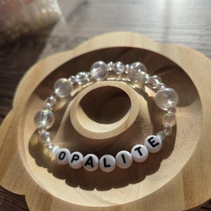 Könnte beinhalten: Ein Armband aus transparenten Perlen mit dem Wort "OPALITE" in weißen Buchstabenperlen. Das Armband liegt auf einem blumenförmigen Holztablett. Die Perlen sind klar und rund.