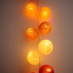 May include: A string of cotton ball lights with warm white and orange bulbs. The lights are strung together with a white wire and are hanging on a white wall.