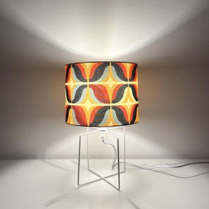 Op de afbeelding: Een retro tafellamp met een cilindrische lampenkap. De lampenkap heeft een herhalend patroon van gebogen vormen in rood, oranje, geel, blauw en zwart. De lamp heeft een witte, geometrische metalen voet en een wit netsnoer.