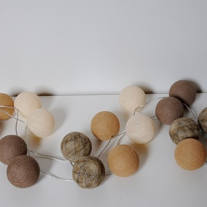 May include: A string of cotton ball lights in beige, brown, and light brown colors. The lights are strung together with a clear wire.