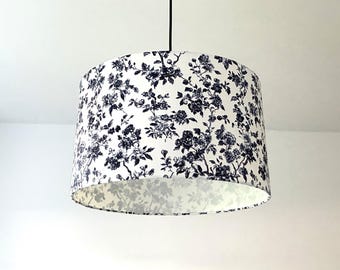 May include: A cylindrical drum lampshade featuring a white background with a dense, repeating dark blue floral pattern. The shade is suspended from a thin black cord, showcasing a classic botanical print that covers the entire exterior surface.