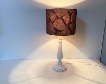 May include: A white table lamp with a dark brown lampshade featuring a geometric pattern. The lamp has a classic, turned base and a cylindrical shade. The lamp is lit, casting a warm glow.