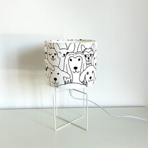 Op de afbeelding: Een tafellamp met een cilindrische witte lampenkap versierd met een zwart lijnpatroon van verschillende hondengezichten. De lamp heeft een eenvoudige witte metalen draadvoet met een kruisvormige onderkant en een wit netsnoer.