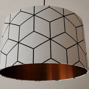 Puede incluir: Una pantalla de lámpara blanca con un patrón geométrico de cubos negros. La parte inferior de la pantalla de la lámpara es de cobre.