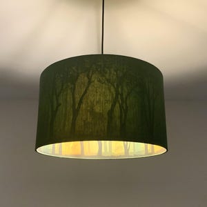 May include: A green lampshade with a forest scene printed on it. The lampshade is lit, and the light shines through the fabric, creating a warm glow.