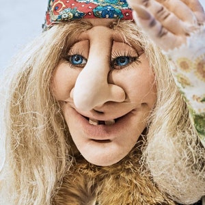 May include: A whimsical doll with long blonde hair, a large nose, and a wide smile. She is wearing a colorful bandana and a fur-trimmed jacket.