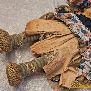 May include: A handmade rag doll with a brown, tan, and white dress, brown crocheted shoes, and a gray and brown striped leg. The doll is lying on a white surface.