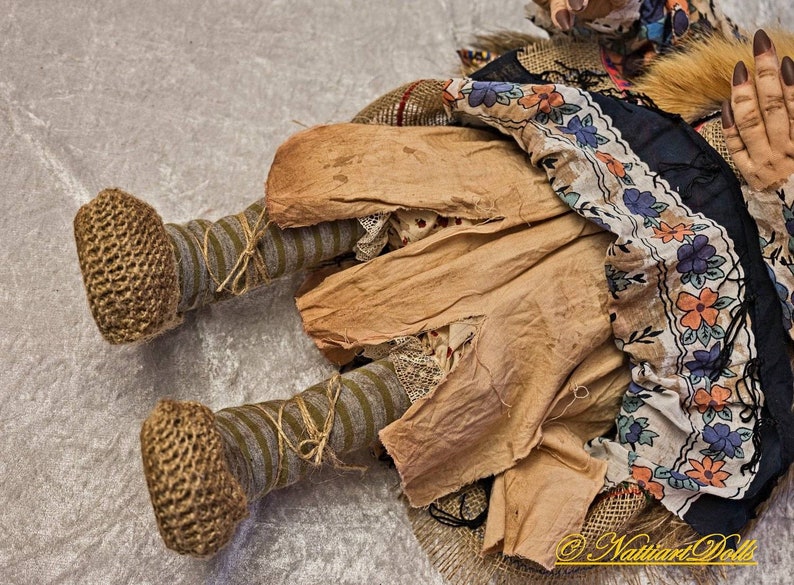 May include: A handmade rag doll with a brown, tan, and white dress, brown crocheted shoes, and a gray and brown striped leg. The doll is lying on a white surface.