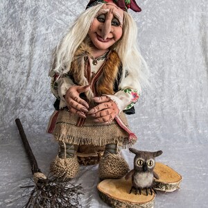 May include: A handmade doll with long white hair, a red and white patterned headscarf, and a brown fur vest. The doll is sitting on a wooden stump and holding a broom. A small brown owl is sitting on a wooden slice next to the doll. The doll is wearing a long brown skirt and white shirt with floral embroidery. The doll is a traditional folk art style.