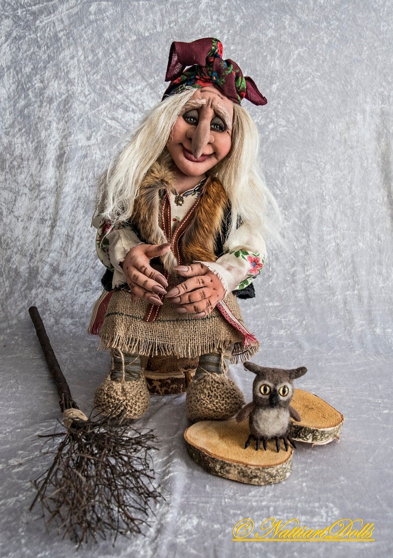 May include: A handmade doll with long white hair, a red and white patterned headscarf, and a brown fur vest. The doll is sitting on a wooden stump and holding a broom. A small brown owl is sitting on a wooden slice next to the doll. The doll is wearing a long brown skirt and white shirt with floral embroidery. The doll is a traditional folk art style.