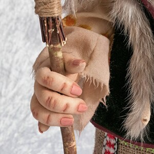 May include: A close-up of a doll's hand holding a wooden stick wrapped in twine. The stick has a frayed end and is wrapped with small twigs. The doll's hand is wearing a tan fabric glove with a frayed edge.