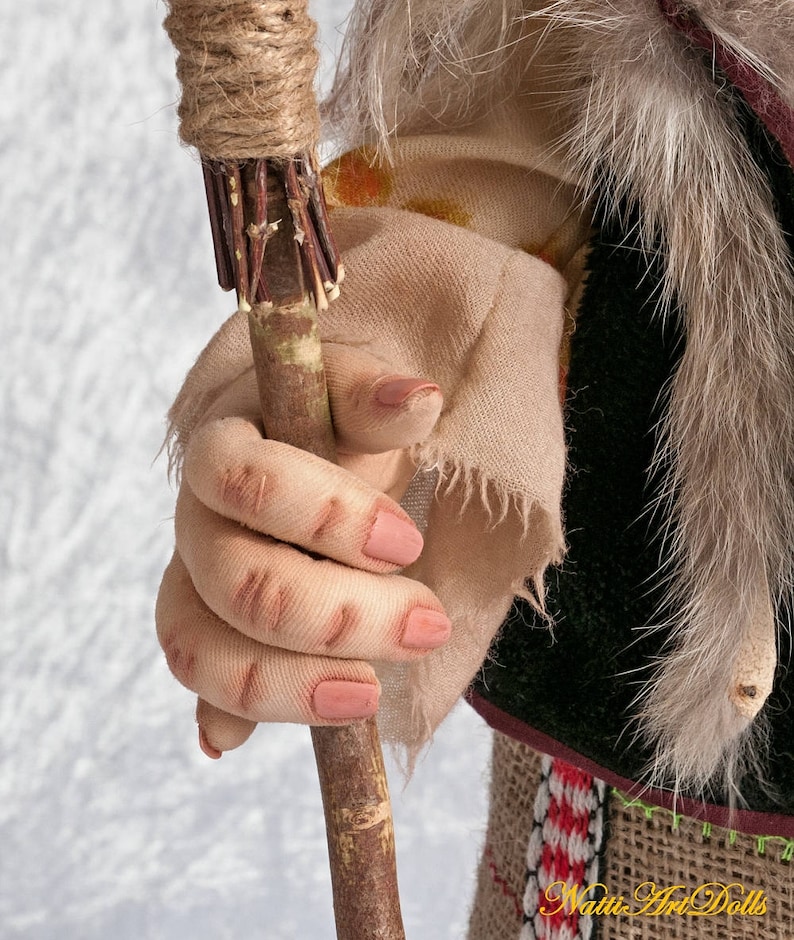 May include: A close-up of a doll's hand holding a wooden stick wrapped in twine. The stick has a frayed end and is wrapped with small twigs. The doll's hand is wearing a tan fabric glove with a frayed edge.