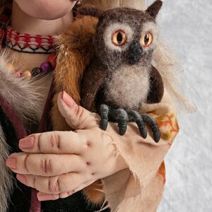 May include: A handmade doll with long blonde hair, wearing a brown and white patterned dress, holds a brown and white owl with yellow eyes. The doll's hand is visible, and the owl's claws are visible.