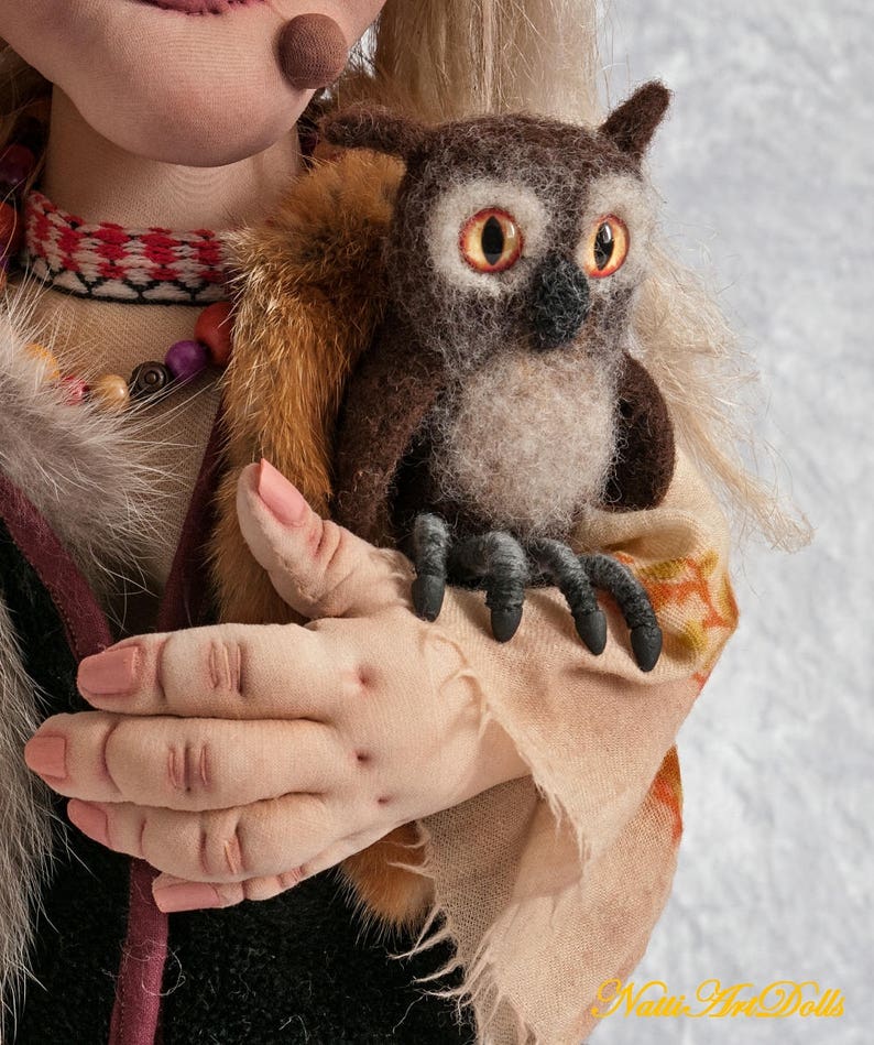 May include: A handmade doll with long blonde hair, wearing a brown and white patterned dress, holds a brown and white owl with yellow eyes. The doll's hand is visible, and the owl's claws are visible.