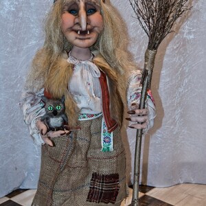 May include: A handmade doll dressed as a witch with a long blonde wig, a red and green bandana, a brown sackcloth skirt, and a brown broom. The doll is holding a small gray stuffed animal with green eyes.