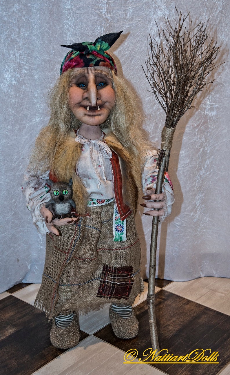 May include: A handmade doll dressed as a witch with a long blonde wig, a red and green bandana, a brown sackcloth skirt, and a brown broom. The doll is holding a small gray stuffed animal with green eyes.