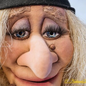 May include: Close-up of a puppet's face with a large, bulbous nose, blue eyes, and a wide smile. The puppet has blonde hair and is wearing a black hat.
