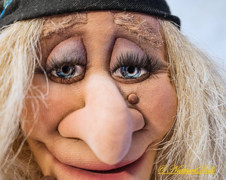 May include: Close-up of a puppet's face with a large, bulbous nose, blue eyes, and a wide smile. The puppet has blonde hair and is wearing a black hat.