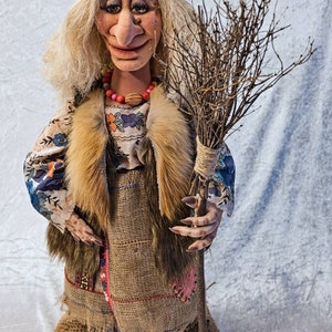 May include: A handmade doll dressed as a witch, with long white hair, a blue and black headscarf, a brown fur vest, a floral print blouse, a brown burlap skirt, and a long twig broom.