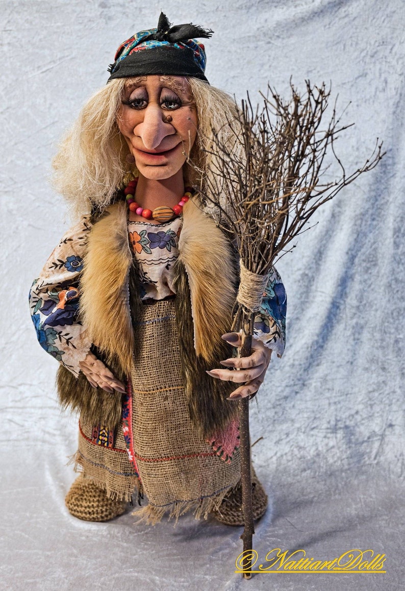 May include: A handmade doll dressed as a witch, with long white hair, a blue and black headscarf, a brown fur vest, a floral print blouse, a brown burlap skirt, and a long twig broom.