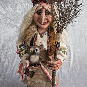 May include: A handmade doll dressed in a traditional folk costume with a red and white patterned skirt, a white blouse with floral embroidery, and a red headscarf. The doll is holding a small owl and a broom made of twigs.