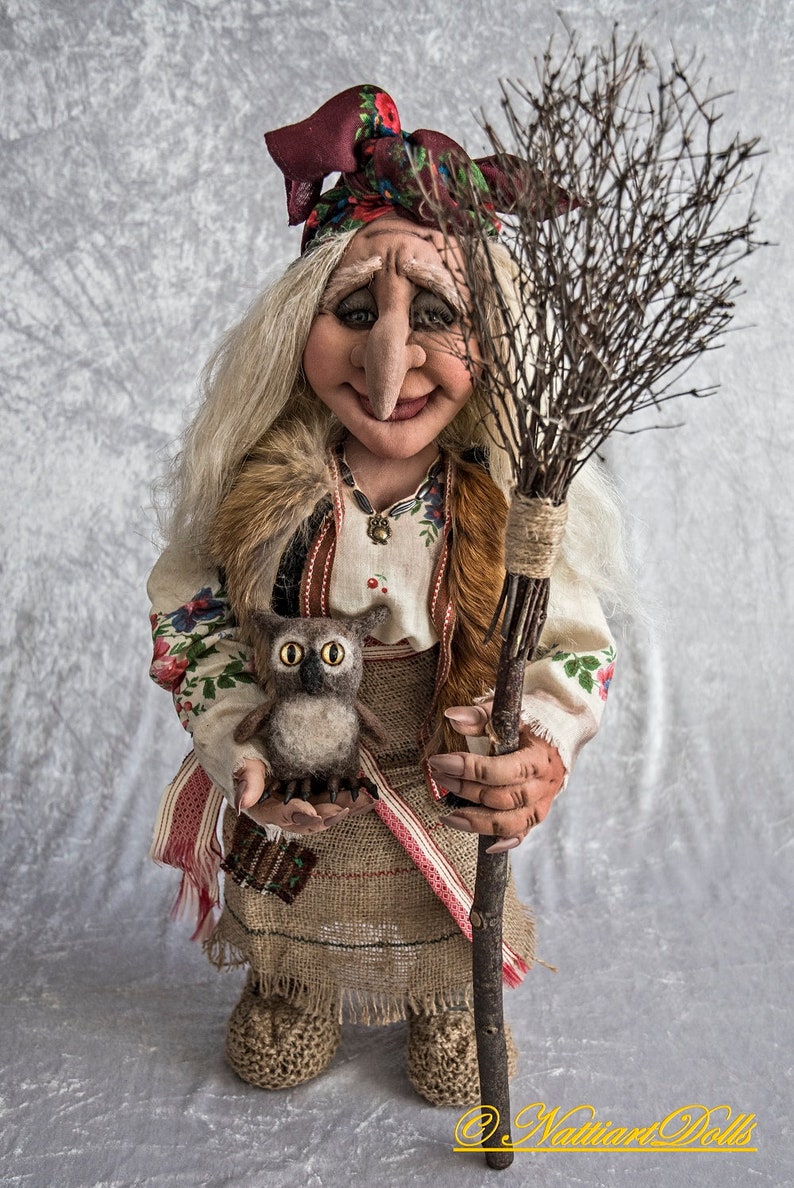 May include: A handmade doll dressed in a traditional folk costume with a red and white patterned skirt, a white blouse with floral embroidery, and a red headscarf. The doll is holding a small owl and a broom made of twigs.
