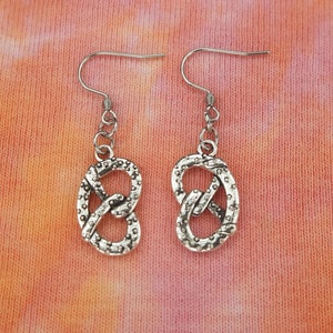 Op de afbeelding: Een paar zilveren oorhangers met een pretzel-ontwerp. De oorbellen zijn op een licht oranje en roze tie-dye achtergrond.