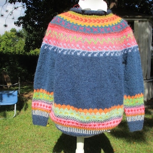 Könnte beinhalten: Ein langärmeliger Strickpullover mit dunkelblauer Basis und einem mehrfarbigen Fair-Isle-Muster an Passe, Ärmelbündchen und Saum. Das Design enthält lebendige horizontale Streifen mit geometrischen Formen in Pink, Orange, Gelb und Hellblau.
