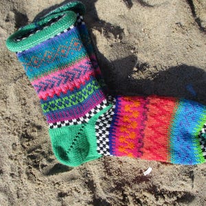 Könnte beinhalten: Eine farbenfrohe Stricksocke mit einem lebendigen Muster aus Rosa, Blau, Grün und Orange. Die Socke hat einen grünen Bund und eine Ferse mit einem schwarz-weißen Karomuster in Knöchelnähe. Die Socke liegt auf sandigem Untergrund.