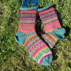 Könnte beinhalten: Ein Paar bunte Strickstrümpfe mit geometrischem Muster. Die Socken haben türkisfarbene Bündchen, Fersen und Zehen sowie Bänder in Orange, Rosa, Grün und Lila. Die Socken liegen auf einer grünen Wiese.