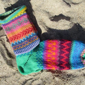 Könnte beinhalten: Eine farbenfrohe Stricksocke mit einem lebendigen Muster aus Pink, Orange, Rot, Blau und Grün. Die Socke liegt auf einer sandigen Oberfläche und zeigt ihr aufwendiges Design und ihre Handwerkskunst. Der Bund der Socke ist ein leuchtendes Gelbgrün.