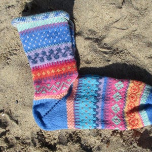 Könnte beinhalten: Eine farbenfrohe Stricksocke mit blauem Bündchen und Zehen sowie einem gemusterten Design in Blau-, Lila-, Rosa-, Orange- und Türkistönen. Die Socke liegt auf einer sandigen Oberfläche.