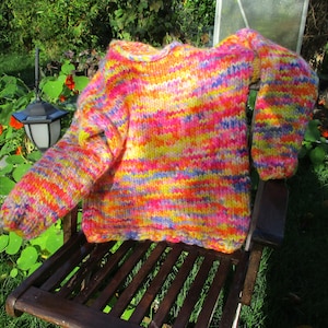 Könnte beinhalten: Ein handgestrickter Pullover in einer lebendigen Farbmischung aus Pink, Gelb, Orange und Blau. Der Pullover wird auf einem Holzstuhl im Freien präsentiert und zeigt sein gemütliches, strukturiertes Aussehen. Die Ärmel sind lang und der Halsausschnitt ist rund.