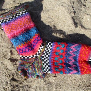 Könnte beinhalten: Eine farbenfrohe, handgestrickte Socke mit einem lebendigen Muster aus rosa, blau, rot und schwarz-weißen geometrischen Designs. Die Socke liegt auf sandigem Untergrund und zeigt ihre komplizierten Details.