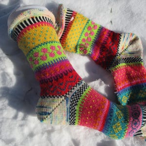 Könnte beinhalten: Ein Paar bunte Strickstrümpfe mit einem lebendigen Muster. Die Socken zeigen Bänder in Gelb, Grün, Rot, Rosa und Blau mit verschiedenen geometrischen und floralen Designs. Die Socken liegen auf einer weißen Schneedecke.